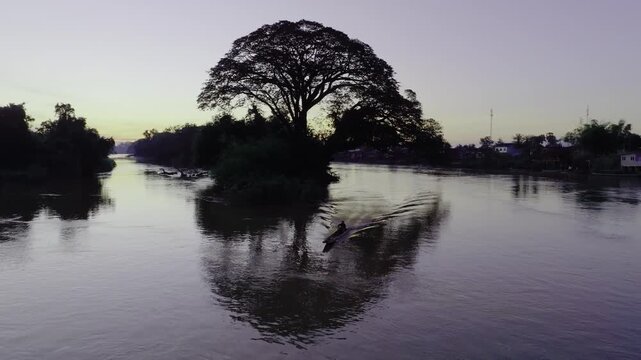 Large tree growing on a small river island at dusk in Don Det, Laos, with a lone boat passing and soft reflections.