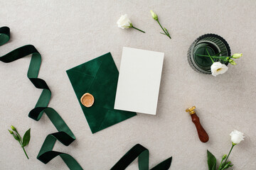 Wedding stationery set. Flat lay wedding invitation card mockup, dark green envelope and ribbon, wax seal stamp, flowers on stone desk table. Top view.