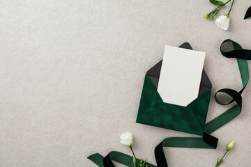 Dark green envelope with wedding invitation card, green ribbon, white flowers on stone background. Top view, flat lay.