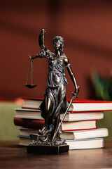 Lady Justice statue holding scales and sword, positioned on a stack of law books with a blurred background of green and brown tones