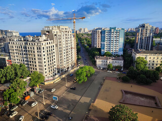 City street view from above. Large multi-story buildings, a square, green trees, an asphalt road for cars, moving cars and walking people on a sunny summer evening