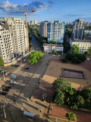 City street view from above. Large multi-story buildings, a square, green trees, an asphalt road for cars, moving cars and walking people on a sunny summer evening