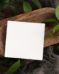 Square card on brown wooden tray near green leaves and grey fabric top view, boho mockup