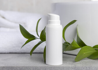 White blank cream bottle near green leaves, basin and white bath towels on grey stone, mockup