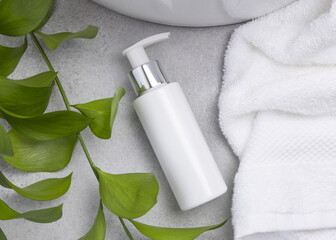 White blank cream bottle near green leaves, basin and white bath towels on grey stone, mockup