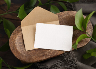 Card and envelope mockup on brown wooden tray near green leaves and grey fabric top view
