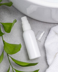 White blank cream bottle near green leaves, basin and white bath towels on grey stone, mockup