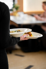 A person in a black uniform is serving two plates of delicious food in a blurred restaurant setting. Focus on the hands and the presented meal