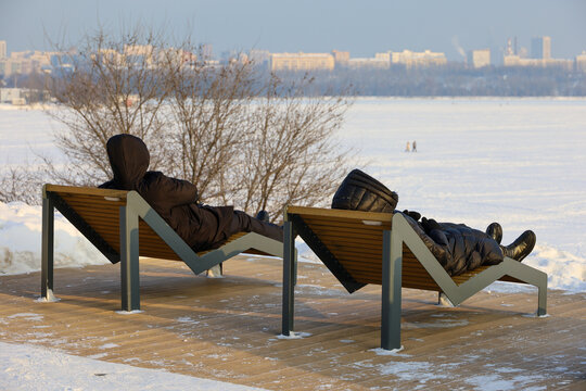 People in warm clothes lying in deck chairs on winter beach on city background. Holidays in cold weather