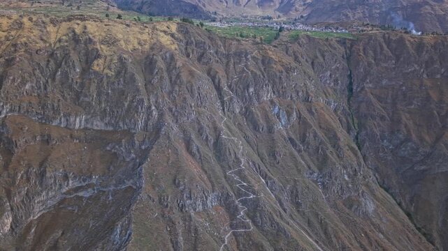 A wide panning drone shot reveals the Colca Canyon&rsquo;s vast scale, showcasing the descending hiking trail and the dramatic contrast between green farmland above and the barren canyon walls below.