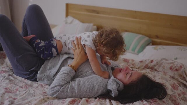 Mother playing with toddler on bed in joyful morning moment as they laugh cuddle lift and lean toward each other creating warm affectionate playful connection 