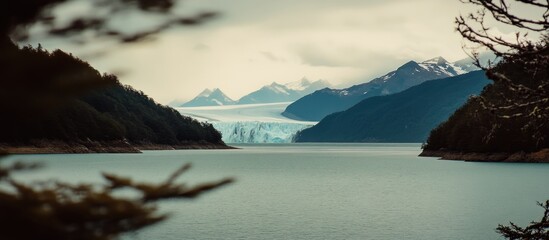 Scenic view of Perito Moreno Glacier in Patagonia, Argentina, featuring a large lake, majestic snow-capped mountains, and lush forests under a cloudy sky, capturing the vast natural landscape.