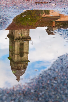 View of the cathedral and tower's reflection on a puddle, with the pebble ground adding texture, creating a surreal and distorted image, Pontremoli, Tuscany, Italy.