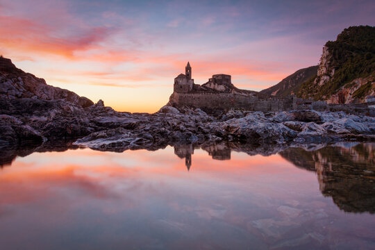 View of a medieval church and castle reflected in the calm waters, under a pastel sunset sky filled with orange and pink hues, Porto Venere, Liguria, Italy.