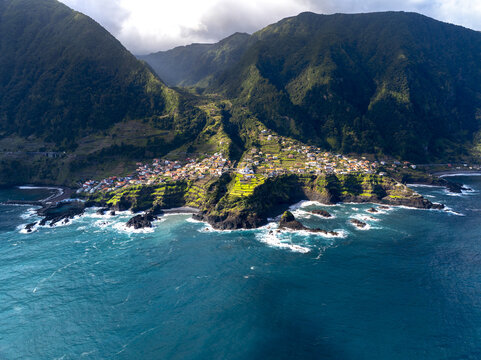 Aerial view of dramatic cliffs meet the turquoise ocean, cradling a village nestled amidst lush green mountains, San vincente, Madeira, Portugal.