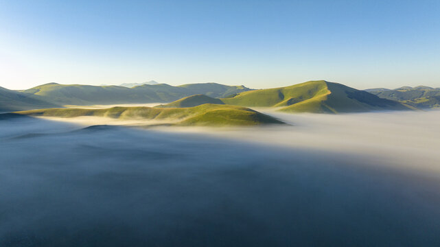 Aerial view of rolling green hills kissed by ethereal white fog, creating a dreamy landscape, Castelluccio, Umbria, Italy.