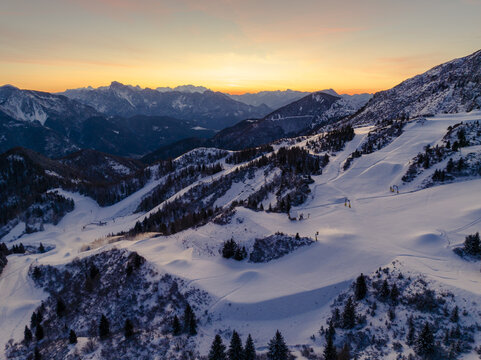 Aerial view of snow-blanketed slopes meeting the rugged peaks under the fiery kiss of sunset, Friuli Venezia Giulia, Italy.