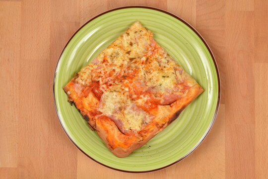 A top down view of a single square slice of homemade pizza with ham and melted cheese served on a light green plate over a wooden surface