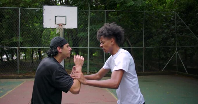 Hispanic and African descent men arguing on outdoor basketball court, one mocking with hand gestures as tension rises and confrontation escalates into physical push