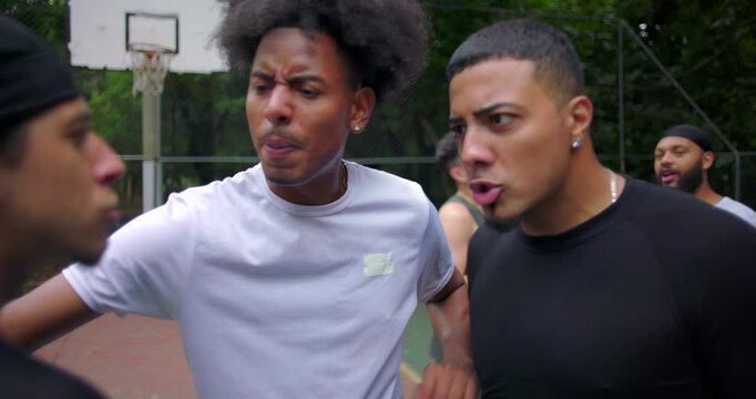 Hispanic men arguing on outdoor basketball court as African descent teammate intervenes to calm conflict, heated confrontation and emotions during street game