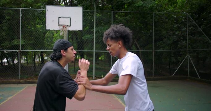 Hispanic and African descent men arguing on outdoor basketball court, one mocking with hand gestures as tension rises and confrontation escalates into physical push