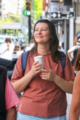 Happy adult woman walking outdoors on a busy city street holding a takeaway coffee cup. Realistic...