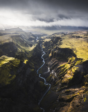 Aerial view of the sinuous river carves through the dark canyon, illuminated by fleeting sunlight under a stormy sky, Sudurland, Iceland.