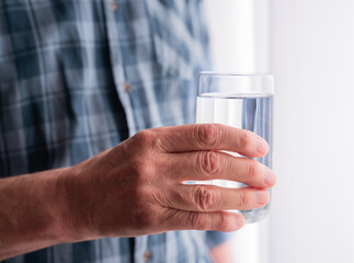Close up of senior man hand holding a glass of water near window in the morning. Healthy lifestyle, hydration habit, daily routine, wellness, calm home atmosphere. Close-up.