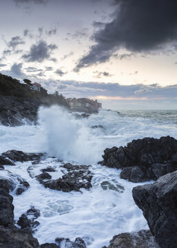 View of frothy waves exploding against dark, jagged rocks under a brooding sky, where the sea meets the shore in Tellaro, Liguria, Italy.