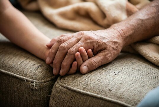 Two hands are clasped together on a couch. One hand is smaller and younger, while the other is larger and older. This scene takes place in a living room, showing a moment of connection