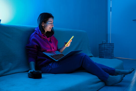 Young woman using smartphone and laptop in blue neon room light