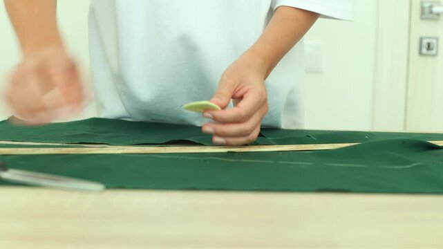 A person in a white shirt uses a wooden ruler and yellow chalk to mark precise lines on dark green fabric, preparing for accurate cutting.