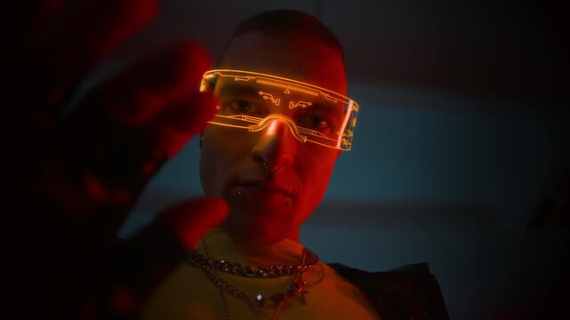 View from behind of young man in futuristic LED glasses with facial piercing interacting with invisible interface in front of camera in dark room