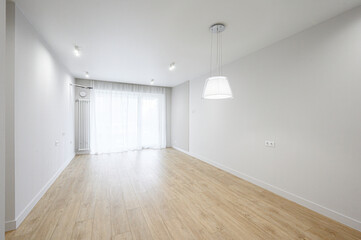 Fototapeta premium A bright, minimalist room with light wooden flooring, white walls, recessed ceiling lights, a pendant lamp, sheer white curtains, and a white radiator — clean, airy space.