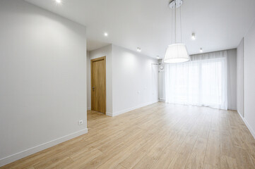 Obraz premium A bright, minimalist room with light wooden flooring, white walls, recessed ceiling lights, a pendant lamp, sheer curtains, and a wooden door — clean, airy, modern space.