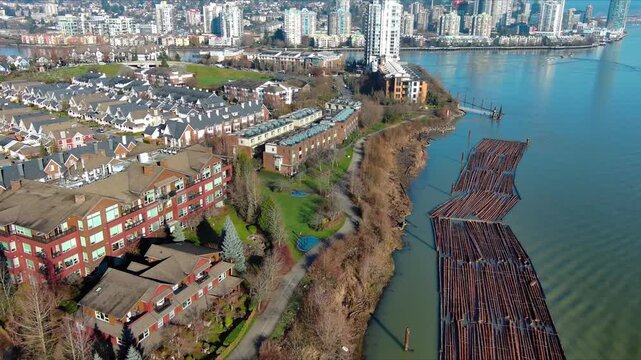 A drone floats above a winding river and log boom. Modern townhomes line the bank, high-rise condos gleam beyond, and snow-capped mountains frame the city at dawn.