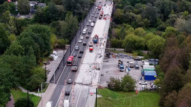 Drone aerial shows highway bridge repair with lane closure and steady traffic beside green parkland, capturing roadworks activity and commuting vehicles in daytime.