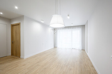 Obraz premium A bright, empty room with light wooden flooring, white walls, recessed ceiling lights, a large pendant lamp, sheer white curtains, and a wooden door — minimalist, airy space.