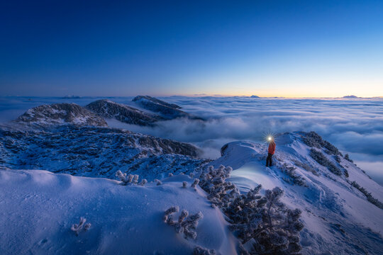 View of a lone figure stands atop a snowy peak, headlamp cutting through the twilight, as a sea of clouds blankets the distant valleys, Srednja vas v Bohinju, Slovenia.
