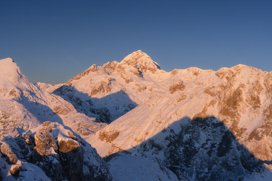 View of snow-dusted mountain peaks bask in the warm glow of the rising sun, casting long shadows across the alpine landscape, Srednja vas v Bohinju, Slovenia.