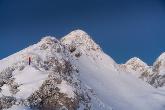 View of a lone figure in a vibrant red jacket stands atop a snow-covered mountain peak against a crisp blue sky, a serene winter vista, Srednja vas v Bohinju, Slovenia.