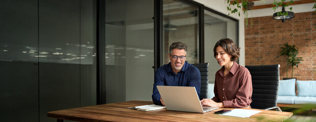 Female manager showing financial results to colleague at work. Two professional team business man and woman using laptop at corporate meeting. Happy busy executive people working together in office.