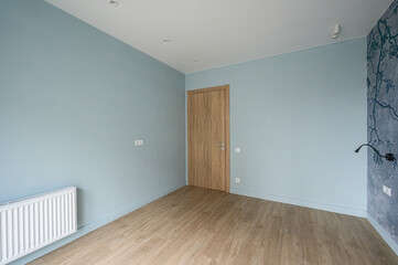 Obraz premium A minimalist room with light blue walls, wooden flooring, wooden door, white radiator, recessed ceiling lights, and a decorative grey wall with a branch pattern — clean, modern space.