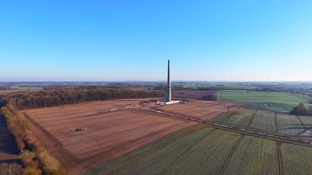 Aerial drone view of a wind turbine tower under construction in a rural agricultural landscape. The footage shows the turbine mast standing in open farmland, surrounded by fields, forests