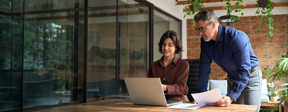 Two busy executive managers working on laptop discussing financial documents at meeting. Middle aged man and woman business professionals team talking planning project at work. Candid photo.