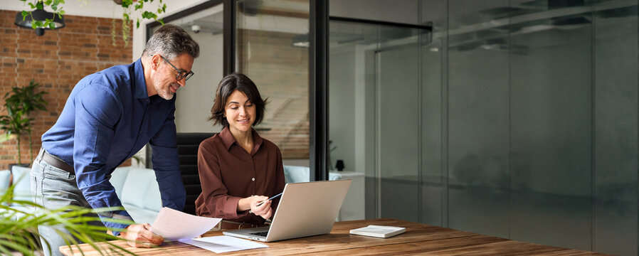 Two happy busy business leaders man and woman talking in office using laptop. Professional executive team people partners checking financial report working together on computer at corporate meeting.