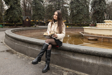 Adult girl is reading a book in a park near a fountain