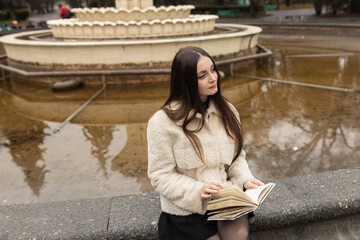 Adult girl is reading a book in a park near a fountain