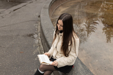 Adult girl is reading a book in a park near a fountain