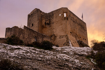 View of the Castle of Vaison-la-Romaine at sunset under a pink sky, with lightly snow-covered ground highlighting the medieval ruins and dramatic winter atmosphere of Provence.

